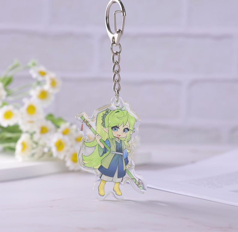 May include: A clear acrylic keychain featuring a cartoon character with green hair and blue eyes, holding a green staff. The character is wearing a blue and green outfit. The keychain has a silver metal chain and clasp.