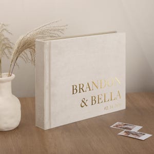 Peut inclure: Un album photo crème avec l'inscription dorée "BRANDON & BELLA" et la date "02.14.2026". L'album est posé sur une surface en bois, avec deux petites photos à proximité. Un vase blanc avec des plantes séchées est en arrière-plan.
