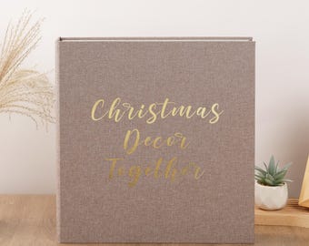 May include: A square photo album with a textured taupe fabric cover. The front features the gold foil text "Christmas Decor Together" in a cursive script. The album has a clean, minimalist design with a sturdy, book-bound construction.