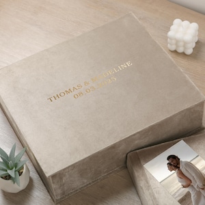 May include: A beige velvet keepsake box with gold lettering reading "THOMAS & MADELINE 08.03.2025." The box is open, revealing a photograph. A small succulent plant in a white pot and a white bubble candle are also visible.