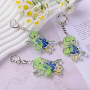 May include: Three clear acrylic keychains featuring a cartoon character with green hair, blue eyes, and a blue and green outfit. Each keychain has a silver-colored chain and clasp. The background includes white flowers and text.