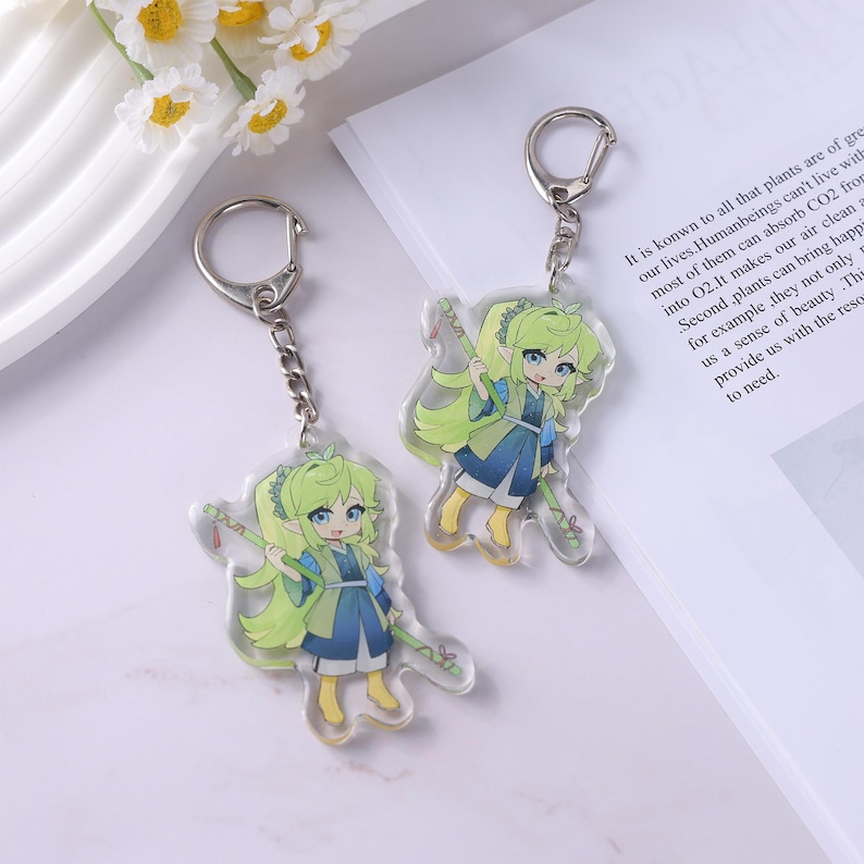 May include: Two clear acrylic keychains featuring a cartoon character with green hair and blue clothing. Each keychain has a silver-colored chain and ring. The character is holding a staff. The background includes text and white flowers.
