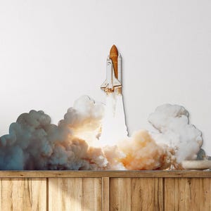 Peut inclure: Un autocollant mural représentant une navette spatiale décollant vers le ciel, entourée d'un grand nuage dense de fumée et de feu blancs et orange. La navette est blanche avec un réservoir externe orange, en position d'ascension verticale.