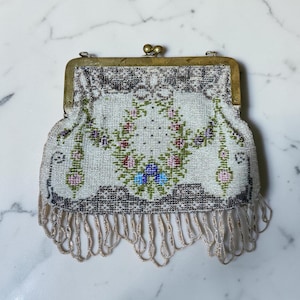 May include: A vintage-style beaded handbag featuring a white background with a floral garland pattern in pink, green, purple, and blue. The bag has a brass-toned metal frame with a double ball clasp and a decorative fringe of hanging beads at the base.