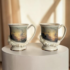 Può includere: Due tazze in ceramica con un design a tramonto. Le tazze hanno una base bianca con bordo e manico dorati. Una decorazione perlata adorna la base di ogni tazza.