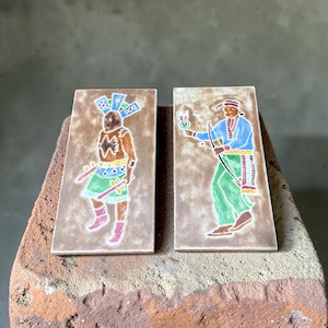 May include: Two rectangular ceramic tiles with a brown background. The left tile features a figure in a blue and green outfit with a feathered headdress. The right tile depicts a figure in a blue shirt and green pants holding a bow and arrow.