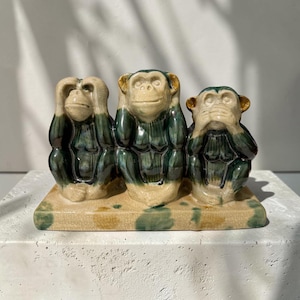 May include: A ceramic figurine featuring three monkeys sitting on a rectangular base. The monkeys are glazed in shades of cream, dark green, and brown, with one covering its eyes, one its ears, and one its mouth, all displaying a crackled finish.