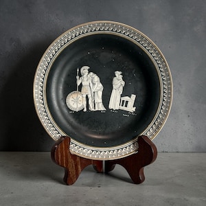 May include: A black plate with a white border and Greek figures. The plate is displayed on a wooden stand. The figures are in white relief against the black background. The border has a repeating wave pattern.