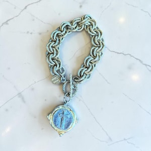 May include: A silver-toned chain link bracelet featuring a toggle clasp and a round pendant with a raised cross design. The metal has a textured, weathered finish. A small circular tag attached to the chain displays the engraved text Susan Shaw.