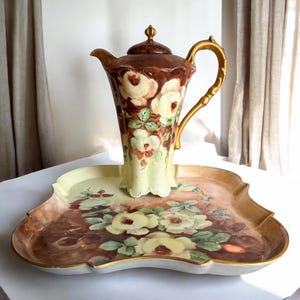 May include: A vintage porcelain coffee pot and serving tray set. The coffee pot is brown with a gold handle and lid, and features a floral design with white and yellow roses. The tray has a matching floral design and gold trim.