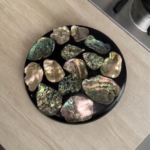 Puede incluir: Una bandeja decorativa redonda y negra llena de conchas de abulón iridiscentes. Las conchas muestran una gama de colores, incluyendo verde, rosa y dorado, creando un efecto brillante. La bandeja está sobre una superficie de madera clara.