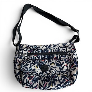 May include: A floral-patterned crossbody bag with a black strap and accents. The bag features a white background with a navy blue floral design, a front pocket, and a zippered main compartment. A black circular logo is visible on the front.