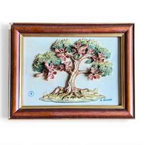 May include: A framed relief sculpture of a tree with green foliage and pink flowers. The tree has a textured, light brown trunk and roots. The frame is a dark brown wood with a gold inner border.