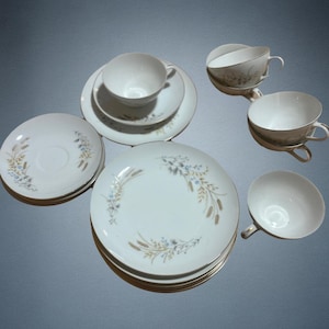 May include: A collection of white porcelain dishware with gold trim. The set includes plates, saucers, and teacups, each adorned with a delicate floral and wheat design in shades of blue and tan. The arrangement suggests a formal dining setting.