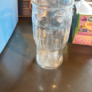 May include: A clear glass tumbler with a face design etched into the glass. The glass has a wide mouth and a slightly tapered base. The design includes eyes, a nose, and a mouth, giving the glass a whimsical appearance. The glass is empty.