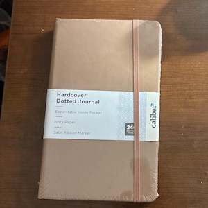 May include: A tan hardcover dotted journal with a matching elastic closure and a satin ribbon marker. The journal has an expandable inside pocket and ivory paper. The label reads "Hardcover Dotted Journal".