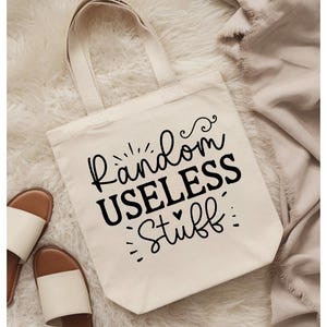 May include: A cream-colored canvas tote bag with two shoulder straps. The front of the bag features black text that reads Random USELESS Stuff, decorated with small flourishes, dots, and a tiny heart symbol centered above the word Stuff.