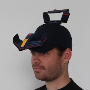 Op de afbeelding: Een donkerblauwe baseballpet met een rood en gele vizierbevestiging. De bevestiging heeft een uniek ontwerp dat lijkt op de voorvleugel van een auto.