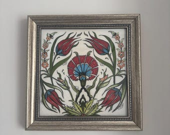 May include: A framed ceramic tile with a hand-painted design of stylized flowers in shades of red, green, blue, and orange. The tile is framed in a silver frame.
