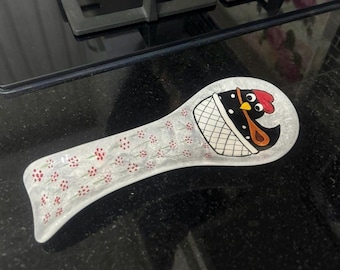 May include: White ceramic spoon rest with a black and red chicken design. The chicken is in a basket with a white and gray pattern. The spoon rest has a white background with small red flowers.