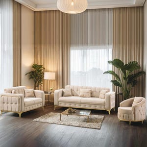 May include: A beige living room suite comprising a sofa, loveseat, and armchair with gold detailing. The furniture is arranged around a glass-topped coffee table on a neutral rug. The room features large windows with beige curtains and indoor plants.