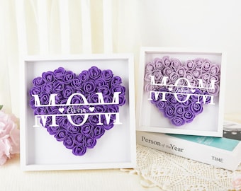 Può includere: Due scatole a ombre incorniciate di bianco con cuori di rose di schiuma viola. I cuori sono disposti a forma della parola "MAMMA" con un nome personalizzato al centro. Il testo "Persona dell'anno" è stampato sul fondo della scatola.