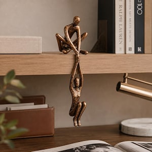 May include: A bronze-colored sculpture featuring two abstract human figures. One figure sits on the edge of a shelf while holding the hands of a second figure that hangs vertically downward, creating a continuous, elongated form with a textured finish.