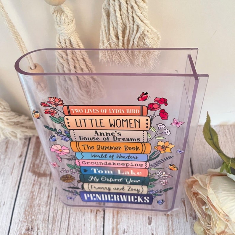 May include: A clear acrylic book stand with a stack of colorful books printed with titles like "Little Women", "Anne's House of Dreams", and "The Summer Book".