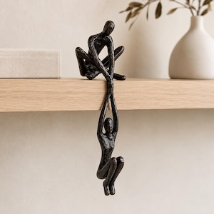 Può includere: Una scultura color bronzo scuro raffigurante due figure umane astratte. Una figura è seduta su una superficie piana e tiene le mani della seconda figura, che pende verticalmente verso il basso dal bordo con le ginocchia ripiegate verso il petto.
