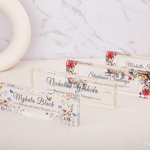 May include: Clear acrylic nameplates with floral designs and names. Each nameplate features a different name, such as Mykala Black, Nicholas Yoshida, Stephanie Santiago, and Michelle Soaknanan, along with the title "Marriage and Family Therapist."