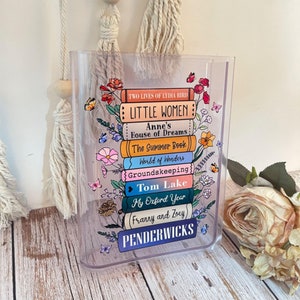 May include: A clear acrylic book-shaped box with a stack of colorful book spines printed on it. The book titles include "Little Women", "Anne's House of Dreams", "The Summer Book", "World of Wonders", "Groundskeeping", "Tom Lake", "My Oxford Year", "(Franny and Zoey)", and "Penderwicks".