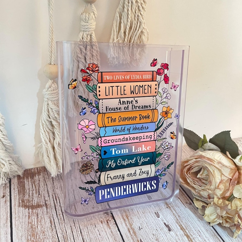 May include: A clear acrylic book-shaped box with a stack of colorful book spines printed on it. The book titles include "Little Women", "Anne's House of Dreams", "The Summer Book", "World of Wonders", "Groundskeeping", "Tom Lake", "My Oxford Year", "(Franny and Zoey)", and "Penderwicks".