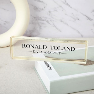 Puede incluir: Una placa de nombre de escritorio de acrílico transparente con el texto "Ronald Toland - Data Analyst" en letras negras.
