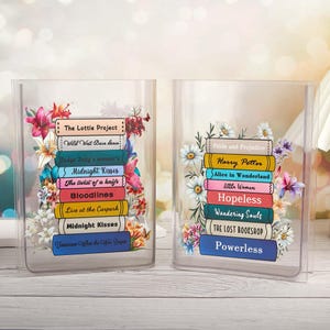 Puede incluir: Dos recipientes transparentes con forma de libro, cada uno con una pila de títulos de libros coloridos y arreglos florales. Los títulos incluyen "The Lottie Project", "Midnight Kisses", "Pride and Prejudice" y "Harry Potter". Los recipientes están decorados con varias flores.