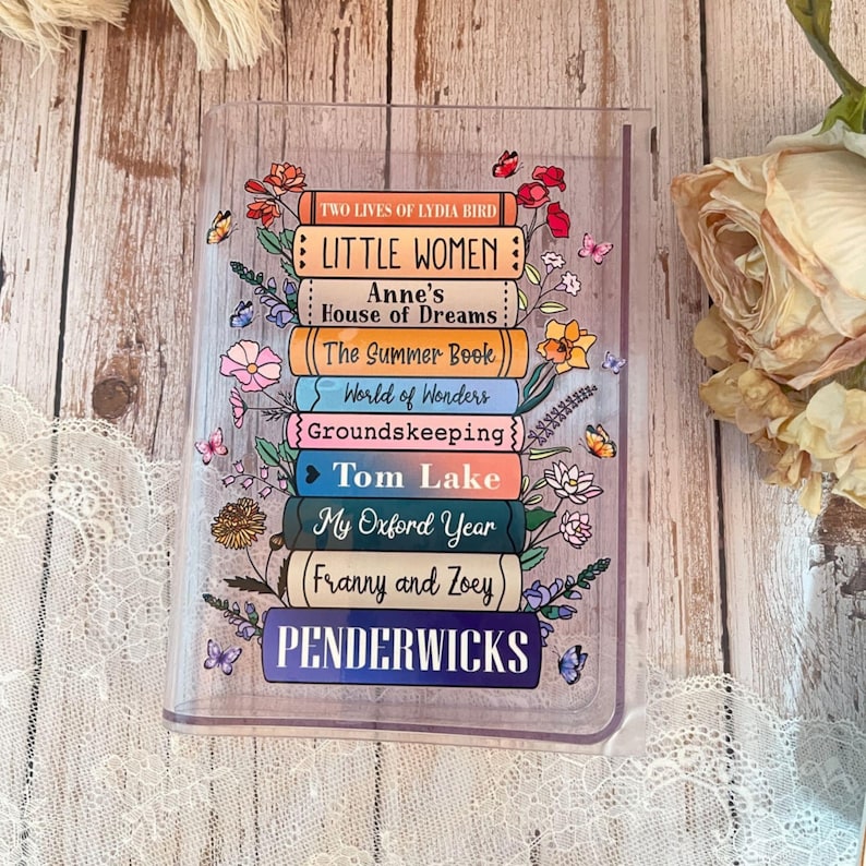 May include: A clear acrylic bookmark with a stack of colorful books with floral accents. The book titles include "Little Women", "Anne's House of Dreams", "The Summer Book", "World of Wonders", "Groundskeeping", "Tom Lake", "My Oxford Year", "Franny and Zoey", and "Penderwicks".