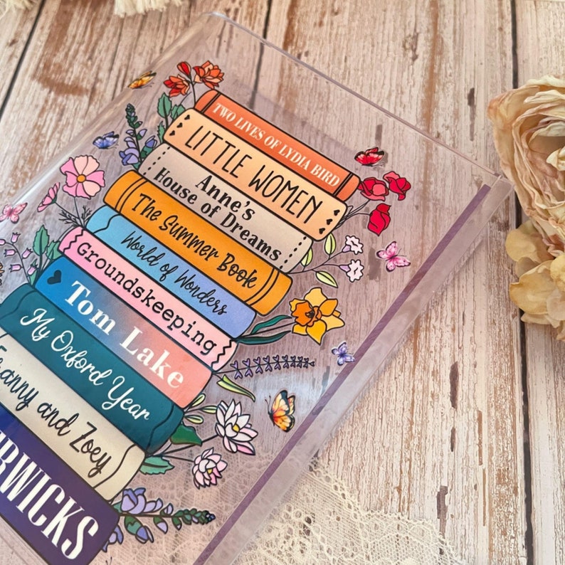 May include: A stack of colorful books with floral illustrations and the titles "Little Women", "Anne's House of Dreams", "The Summer Book", "World of Wonders", "Groundskeeping", "Tom Lake", "My Oxford Year", and "aury and Zoey" visible.