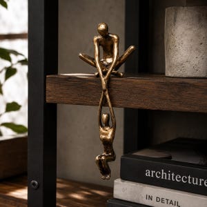 May include: A bronze-toned metal sculpture featuring two abstract figures. One figure sits on the edge of a shelf with legs crossed, while the other figure hangs vertically below, held by the upper figure's hand, creating a connected vertical form.