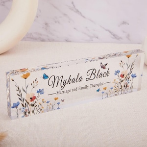 May include: A clear acrylic block featuring the name "Mykala Black" in elegant script, with the title "Marriage and Family Therapist" below. The block is adorned with a floral design in shades of blue, orange, and red, and includes butterfly accents.
