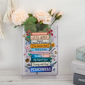 May include: A clear acrylic vase decorated with a stack of book titles, including "Little Women" and "Tom Lake." The vase holds pink flowers and greenery. Butterflies and floral accents surround the book titles, creating a literary and decorative theme.