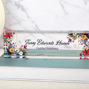 May include: A clear acrylic desk nameplate with floral accents. The nameplate features the name "Tansy Edwards Hamm" and the title "Certified Nutritionist" in elegant script. Colourful flowers in red, yellow, and blue adorn the corners.
