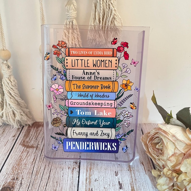 May include: A clear plastic pencil holder with a stack of colorful books printed on the front. The books are decorated with flowers and butterflies. The titles of the books are "Two Lives of Lydia Bird", "Little Women", "Anne's House of Dreams", "The Summer Book", "World of Wonders", "Groundskeeping", "Tom Lake", "My Oxford Year", "(Franny and Zoey)", and "Penderwicks".