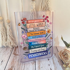 May include: A clear acrylic bookend with a stack of colorful books printed with the titles "Little Women", "Anne's House of Dreams", "The Summer Book", "World of Wonders", "Groundskeeping", "Tom Lake", "My Oxford Year", "Franny and Zoey", and "Penderwicks". The bookend is decorated with floral illustrations.