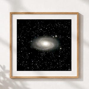 May include: A square framed wall art print featuring a black and white depiction of a spiral galaxy centered against a dark starry background. The artwork is encased in a light-toned wooden frame with a white mat border surrounding the image.
