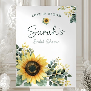 May include: A white sign with the text "Love in Bloom" and "Sarah's Bridal Shower." The sign features a large yellow sunflower and green foliage. The design is a watercolor style, with soft edges and a delicate aesthetic.