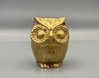 May include: A gold-colored owl figurine. The owl has large, detailed eyes and a textured surface. The figurine is a decorative item, likely for home decor or as a collectible. The owl is standing upright.