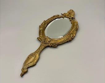 May include: A gold-toned hand mirror with an ornate, detailed frame and a handle. The mirror is oval-shaped and has a clear glass surface.