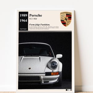Puede incluir: Un póster enmarcado con un Porsche 911 964 blanco. El póster incluye el logotipo de Porsche y el texto: "1989 Porsche 911 964 1964 Sportwagen Form folgt Funktion." La parte delantera del coche es visible sobre un fondo oscuro.
