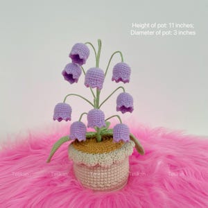 May include: A handcrafted crochet flower arrangement featuring lavender bell-shaped flowers. The plant is in a beige pot with a scalloped edge, sitting on a pink surface. The pot is 11 inches high with a 3-inch diameter.