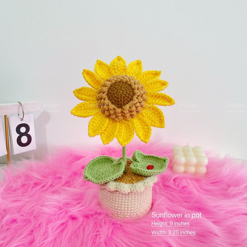 May include: A handmade crochet sunflower in a pot. The sunflower has bright yellow petals and a brown center. The pot is beige with a scalloped edge. The height is 9 inches and the width is 3.25 inches.