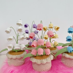 May include: Crocheted flower arrangements in various colors, including white, pink, yellow, purple, and blue. Each flower is in a small, beige crocheted pot with green leaves. The flowers are arranged on a pink surface.